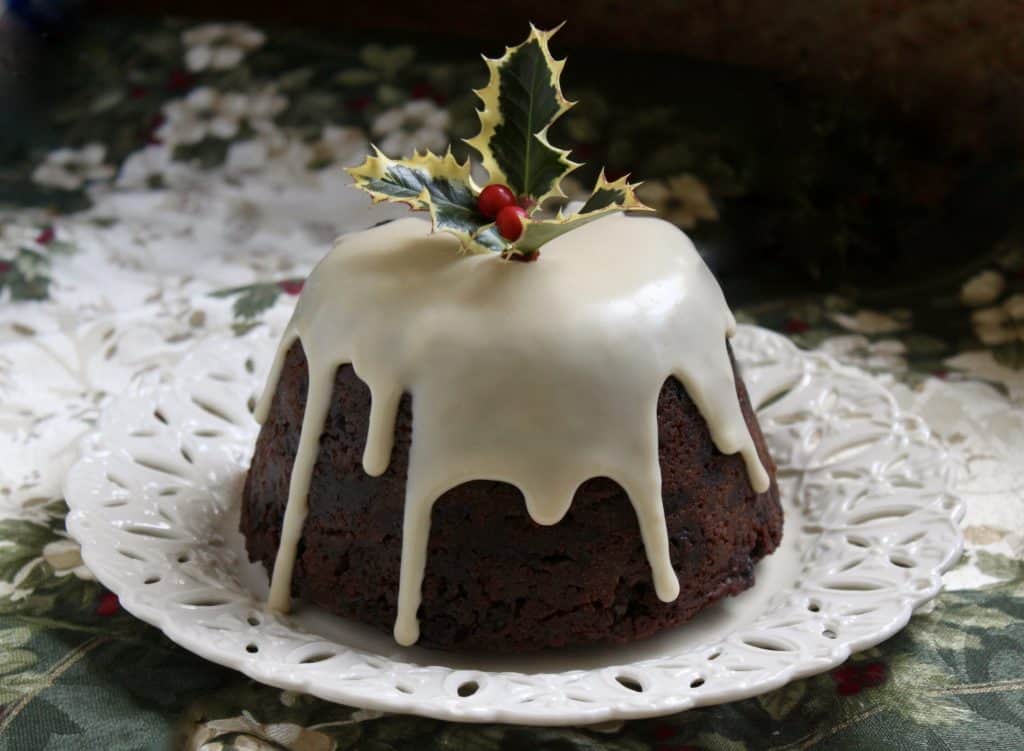 A round fruitcake with white icing dripping down the sides, topped with a sprig of holly, sits on a white decorative plate.