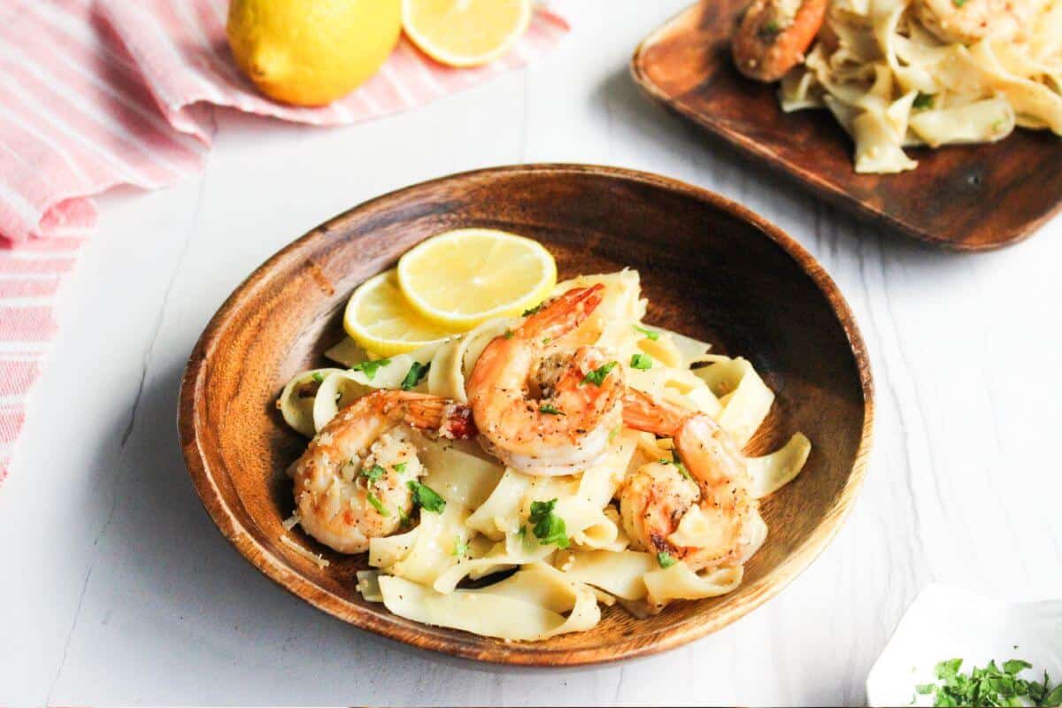 A wooden bowl filled with shrimp and pasta garnished with parsley and lemon slices sits on a white table.