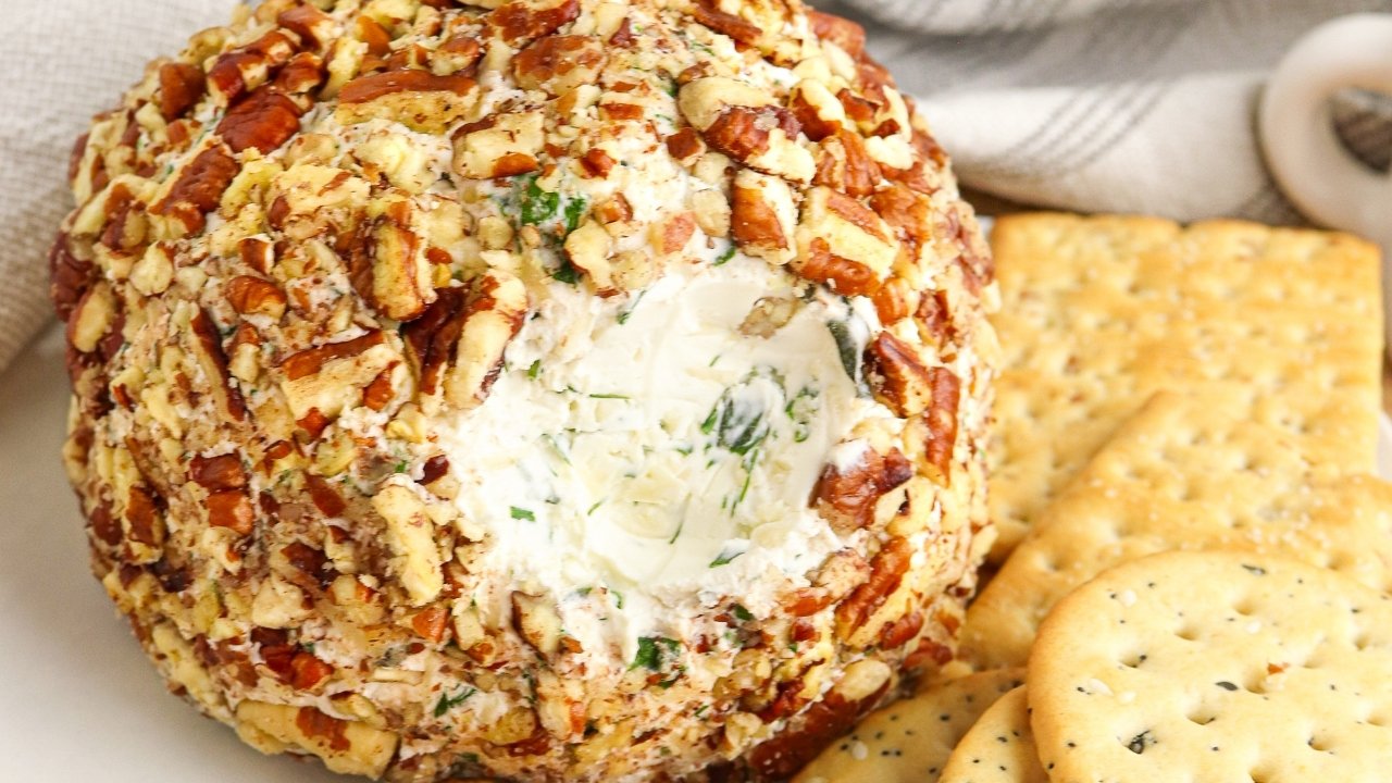 A cheese ball coated in chopped nuts sits on a plate next to round and square crackers. A portion of the cheese ball has been scooped out, revealing a creamy interior with herbs.