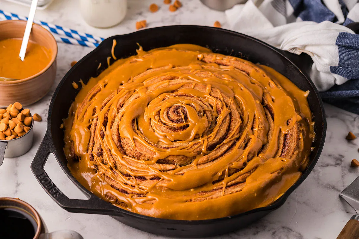 A large cinnamon roll topped with caramel icing sits in a black skillet on a marble countertop, surrounded by baking ingredients and utensils.