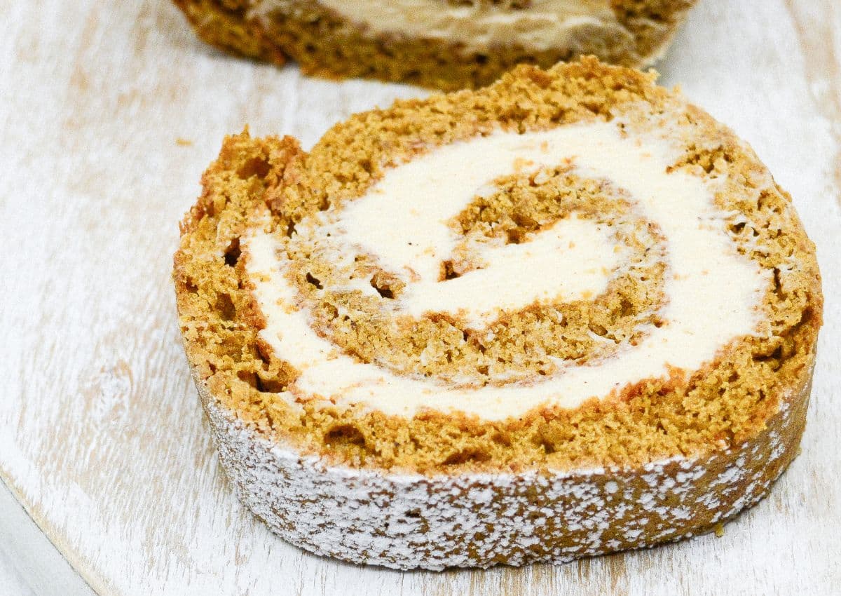 A close-up of a slice of pumpkin roll cake with cream cheese filling on a light wooden surface, dusted with powdered sugar.