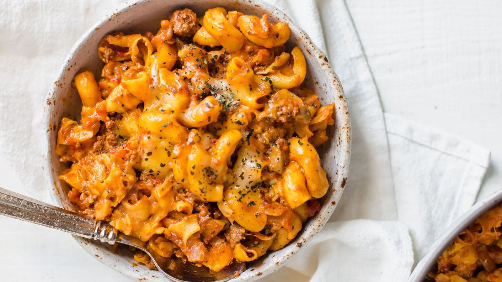 A bowl of cheesy pasta with ground meat and tomato sauce, topped with herbs, on a white cloth with a spoon.