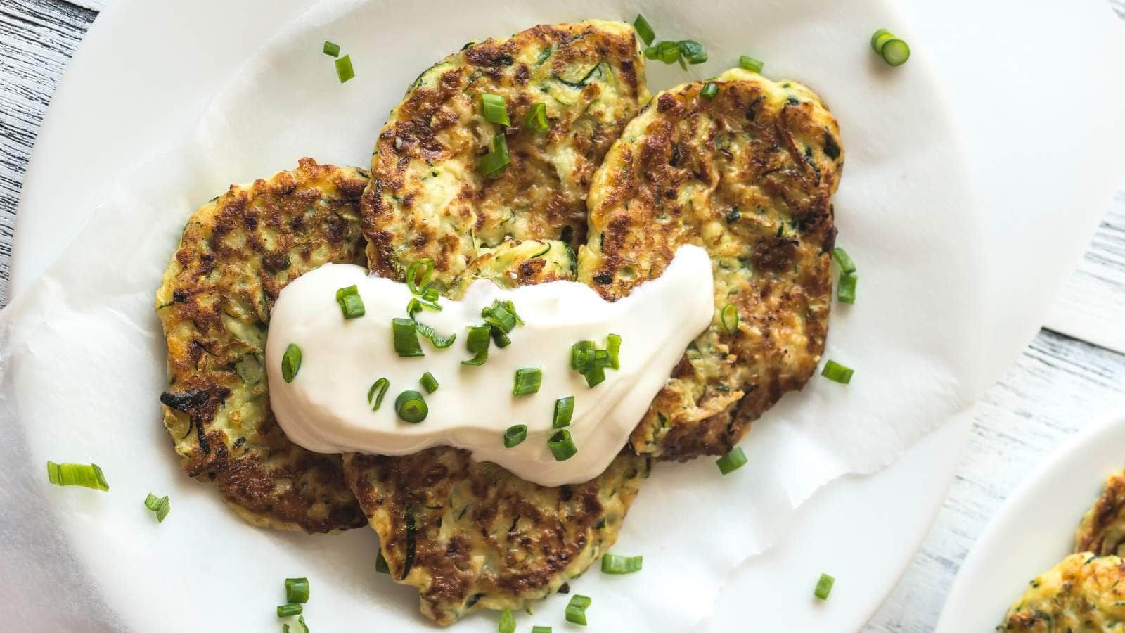 Three zucchini fritters on a white plate, topped with a dollop of sour cream and sprinkled with chopped green onions.