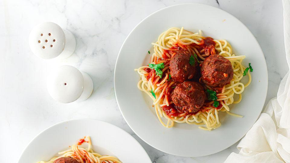 A plate of spaghetti topped with tomato sauce, three meatballs, and parsley, next to another partial plate and salt and pepper shakers on a white surface.