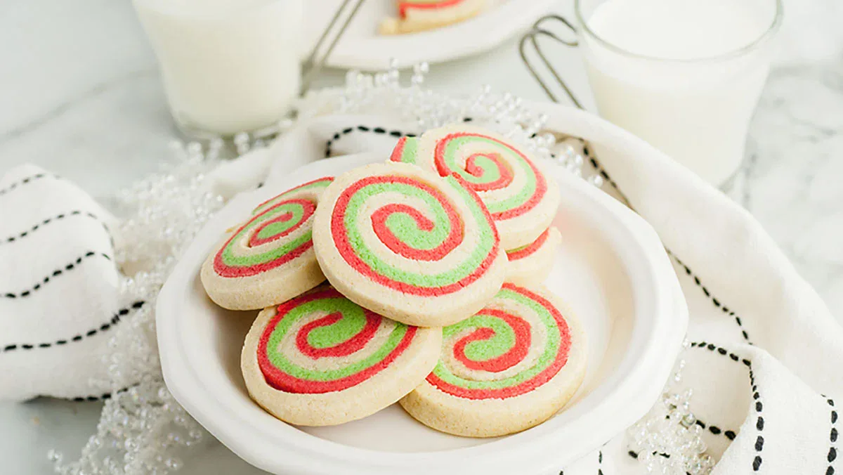A plate of spiral sugar cookies with red and green swirls sits on a white napkin, with glasses of milk in the background.