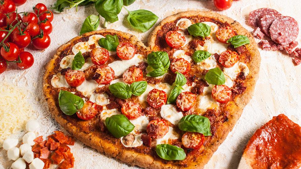 Heart-shaped pizza topped with cherry tomatoes, mozzarella, and basil, surrounded by fresh ingredients like cheese, tomatoes, basil, salami, and pizza dough on a floured surface.
