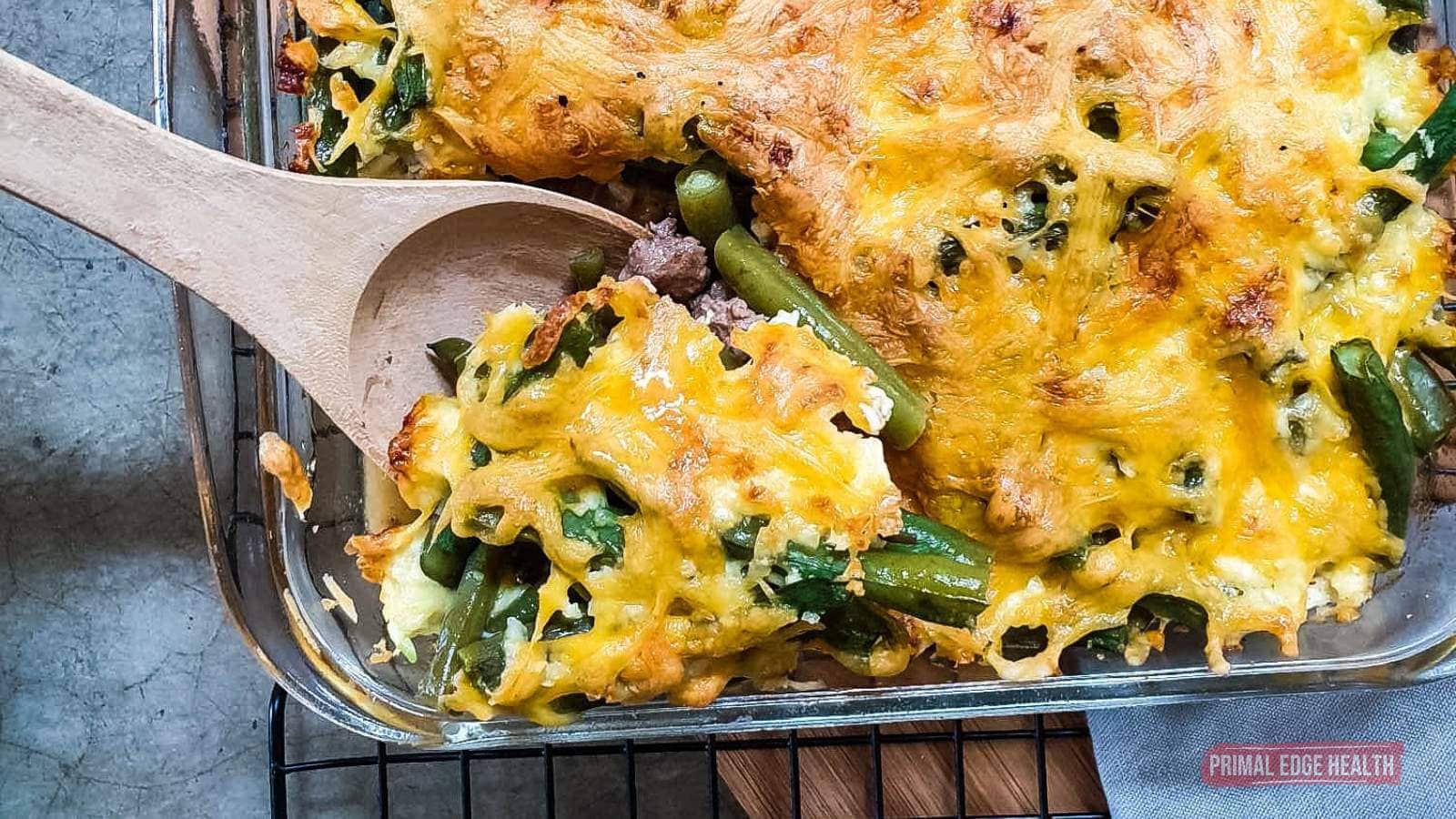 A baked casserole topped with melted cheese, green beans, and other vegetables, with a wooden spoon lifting a serving from a glass baking dish.