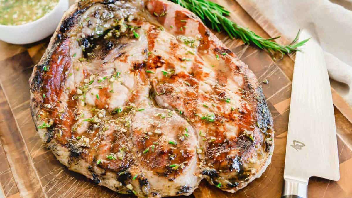Grilled ham steak garnished with herbs on a wooden cutting board, next to a knife and a sprig of rosemary.