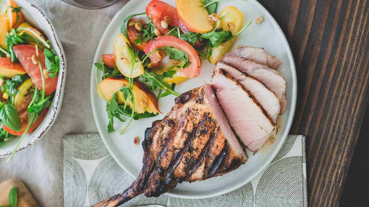 A grilled bone-in pork chop, sliced, served with a salad of tomatoes, peaches, arugula, and pine nuts on a white plate.