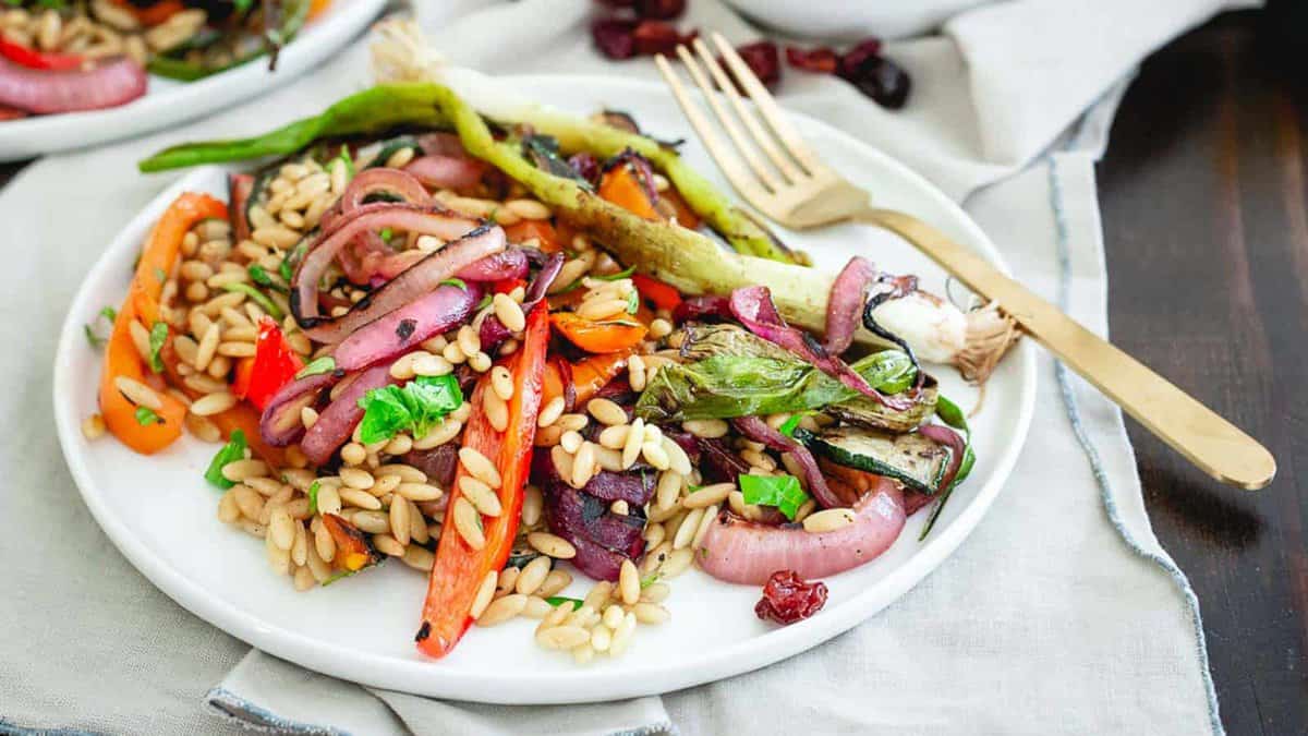 A white plate with cooked orzo pasta, grilled vegetables such as bell peppers, onions, and green beans, garnished with fresh herbs. A gold fork rests beside the plate.