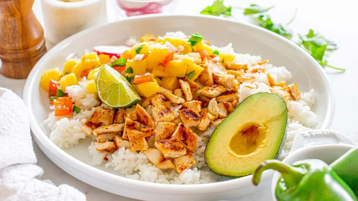 A plate of white rice topped with diced grilled chicken, mango salsa, half an avocado, and a lime wedge, garnished with herbs.
