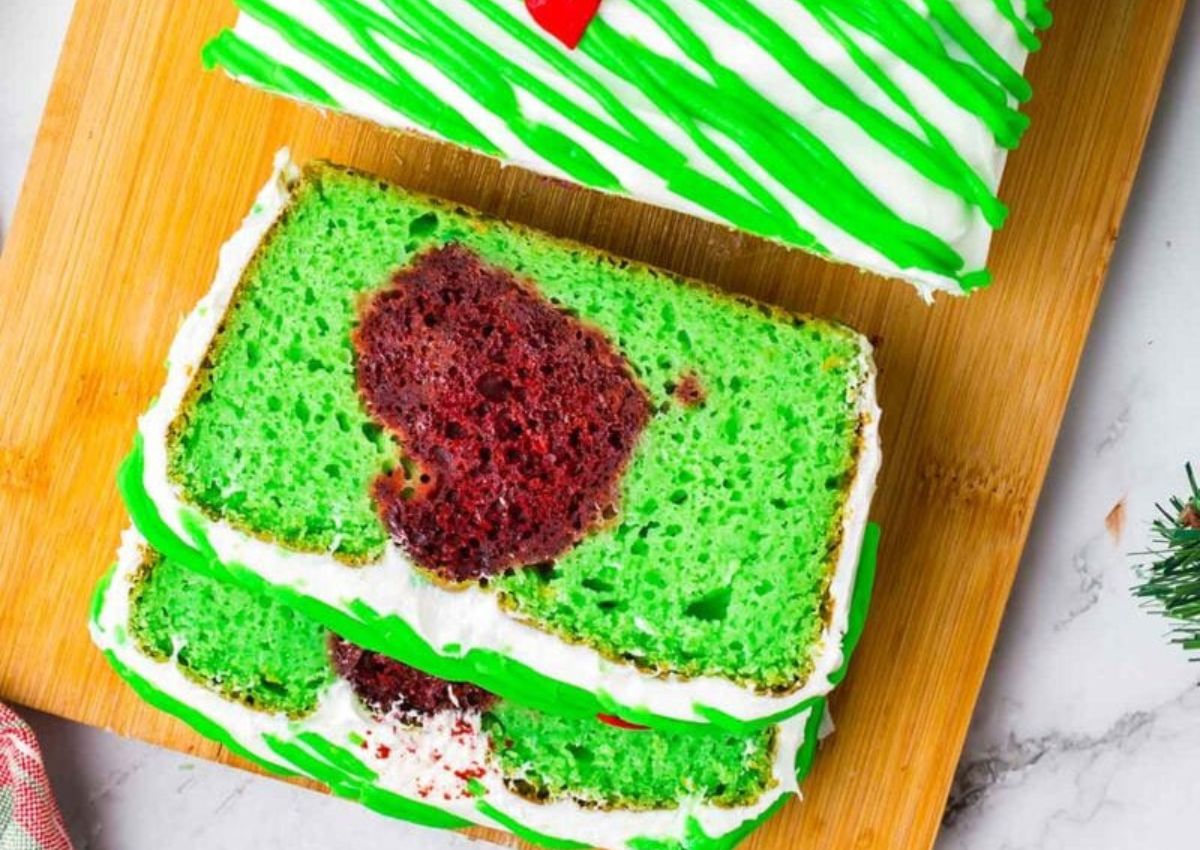 Sliced green loaf cake with white and green icing, revealing a red heart-shaped design in the center, displayed on a wooden cutting board.