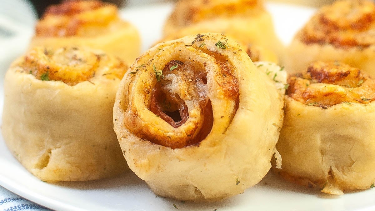 Close-up of several golden-brown pizza pinwheels on a white plate, showing rolled dough with visible cheese and tomato filling.