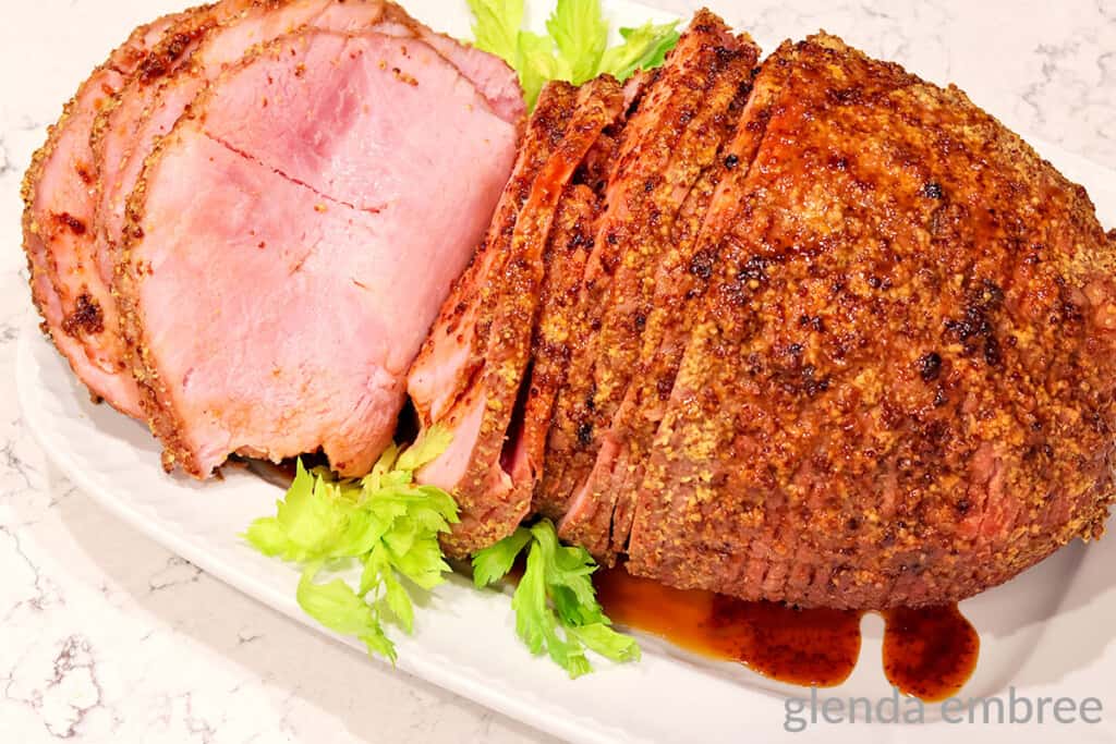 Sliced, seasoned baked ham on a white platter, garnished with celery leaves and a small pool of juices.