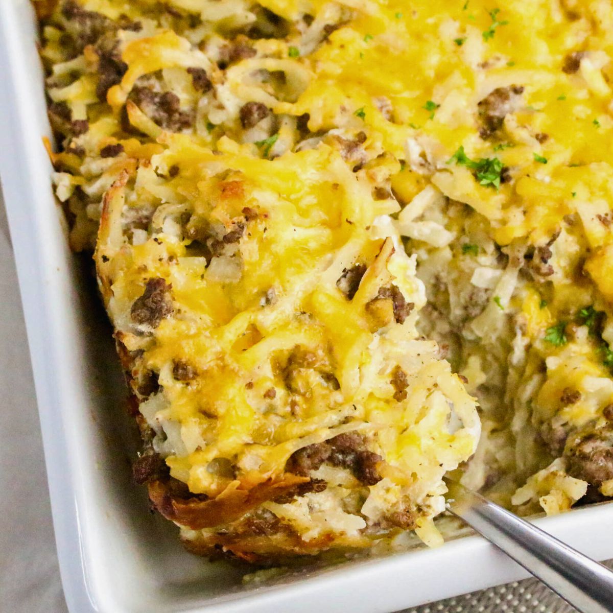 A close-up of a baked casserole with ground beef, shredded potatoes, and melted cheese in a white dish, with a serving being lifted out.