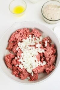 A bowl with raw ground beef and chopped onions, surrounded by small bowls containing melted butter, a creamy mixture, and other ingredients.