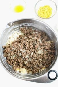 A mixing bowl with cooked ground beef and onions on top of shredded hash browns, with small bowls of shredded cheese and broth nearby.