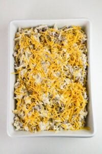 Rectangular white baking dish filled with a mixture of shredded cheese, hash browns, and sausage, topped with more shredded cheese, on a white surface.