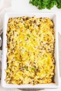 A baked casserole in a white dish, topped with melted cheese, hash browns, and ground meat, with fresh parsley garnish nearby.