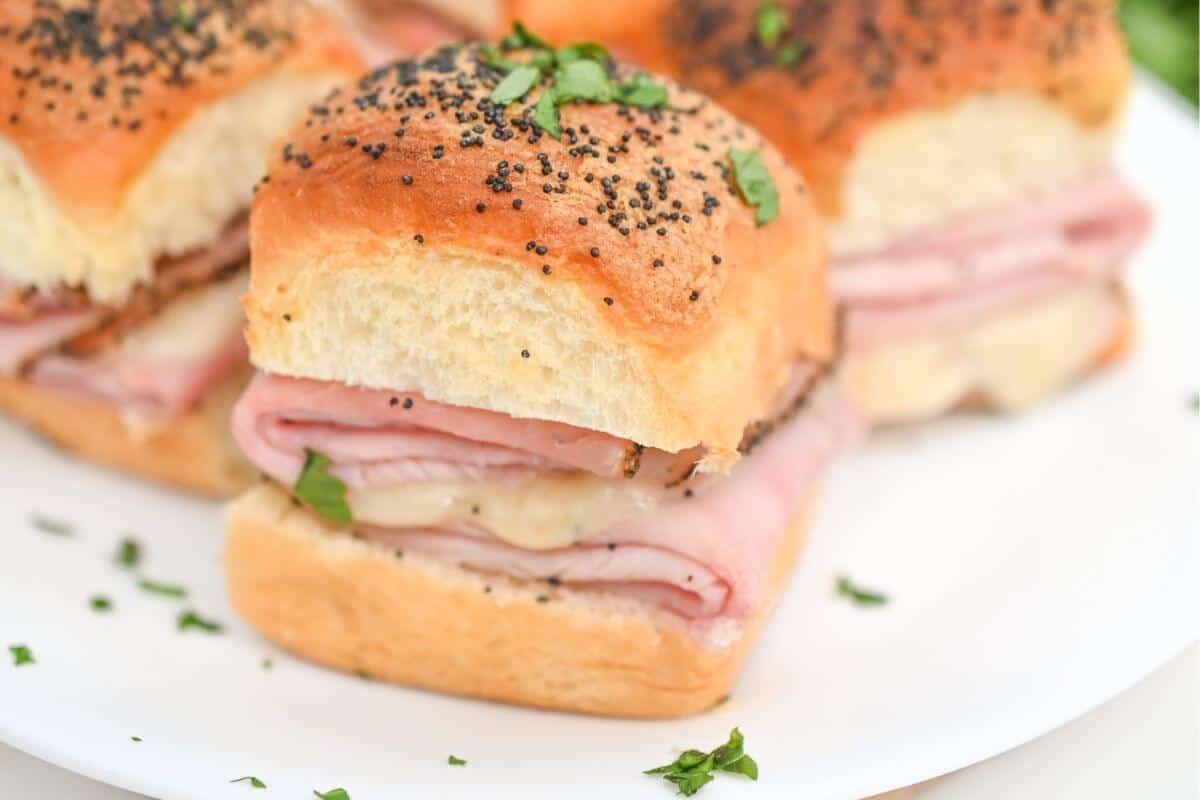 Close-up of ham and cheese sliders on poppy seed buns, garnished with chopped parsley, served on a white plate.