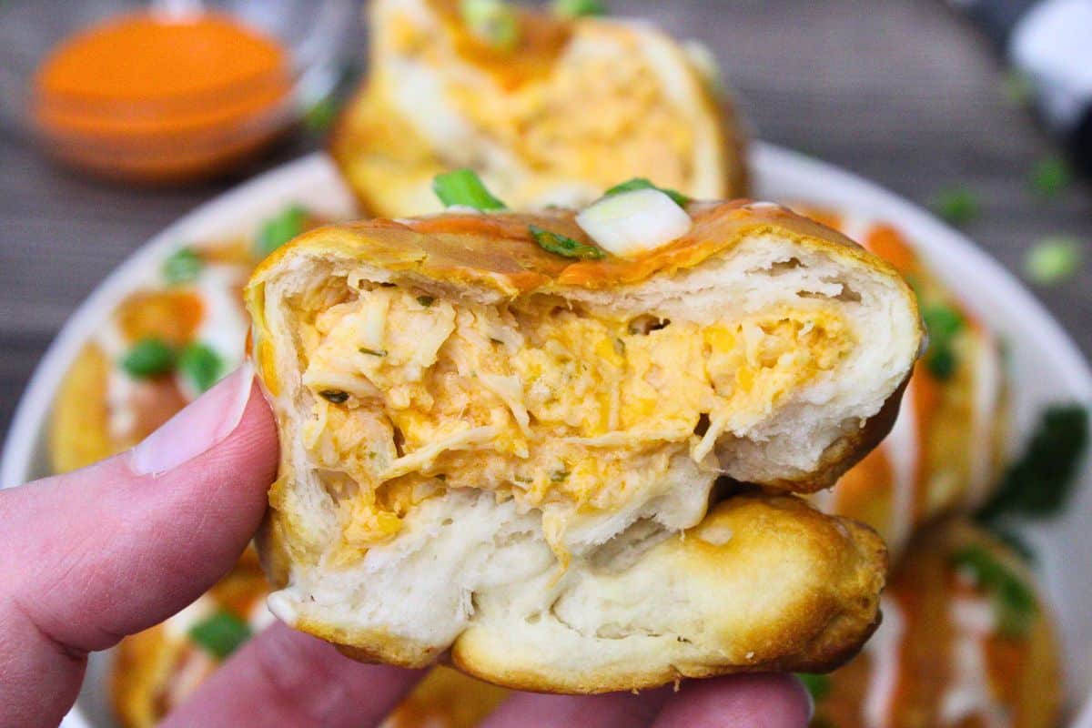 A hand holds a partially eaten stuffed bread roll filled with creamy, shredded chicken. More stuffed rolls with orange sauce and chopped green onions are visible in the background.