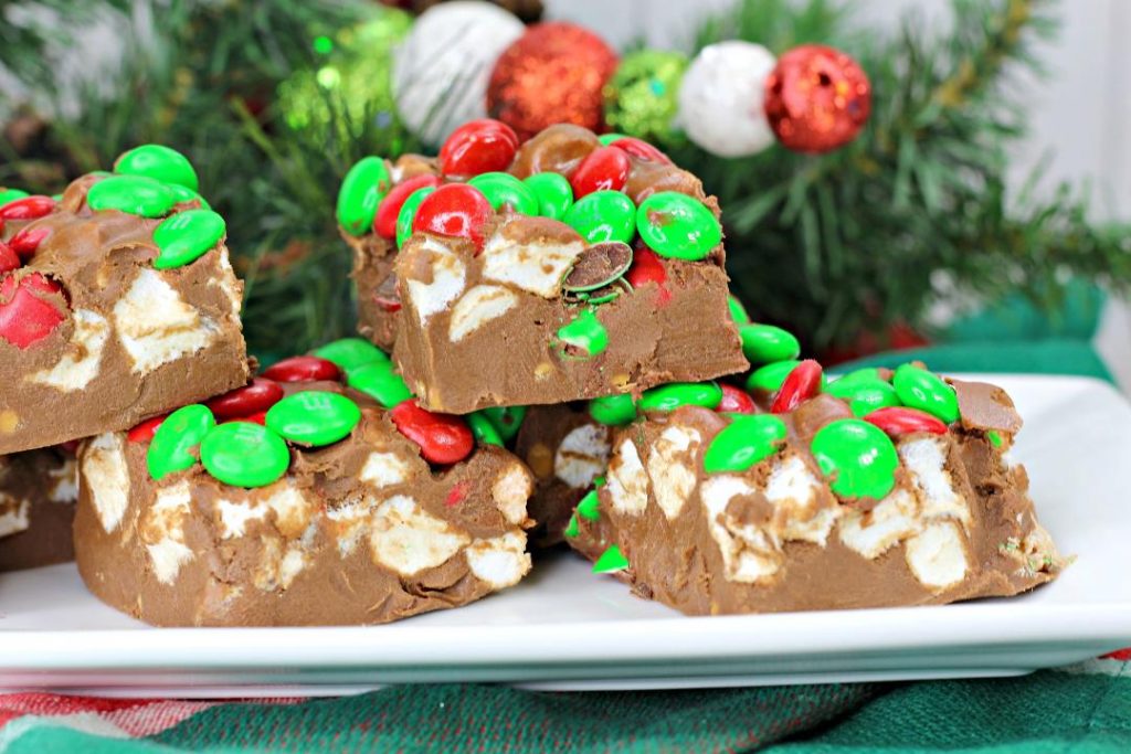 Squares of chocolate fudge with mini marshmallows and red and green candy-coated chocolates, displayed on a white plate with holiday decorations in the background.