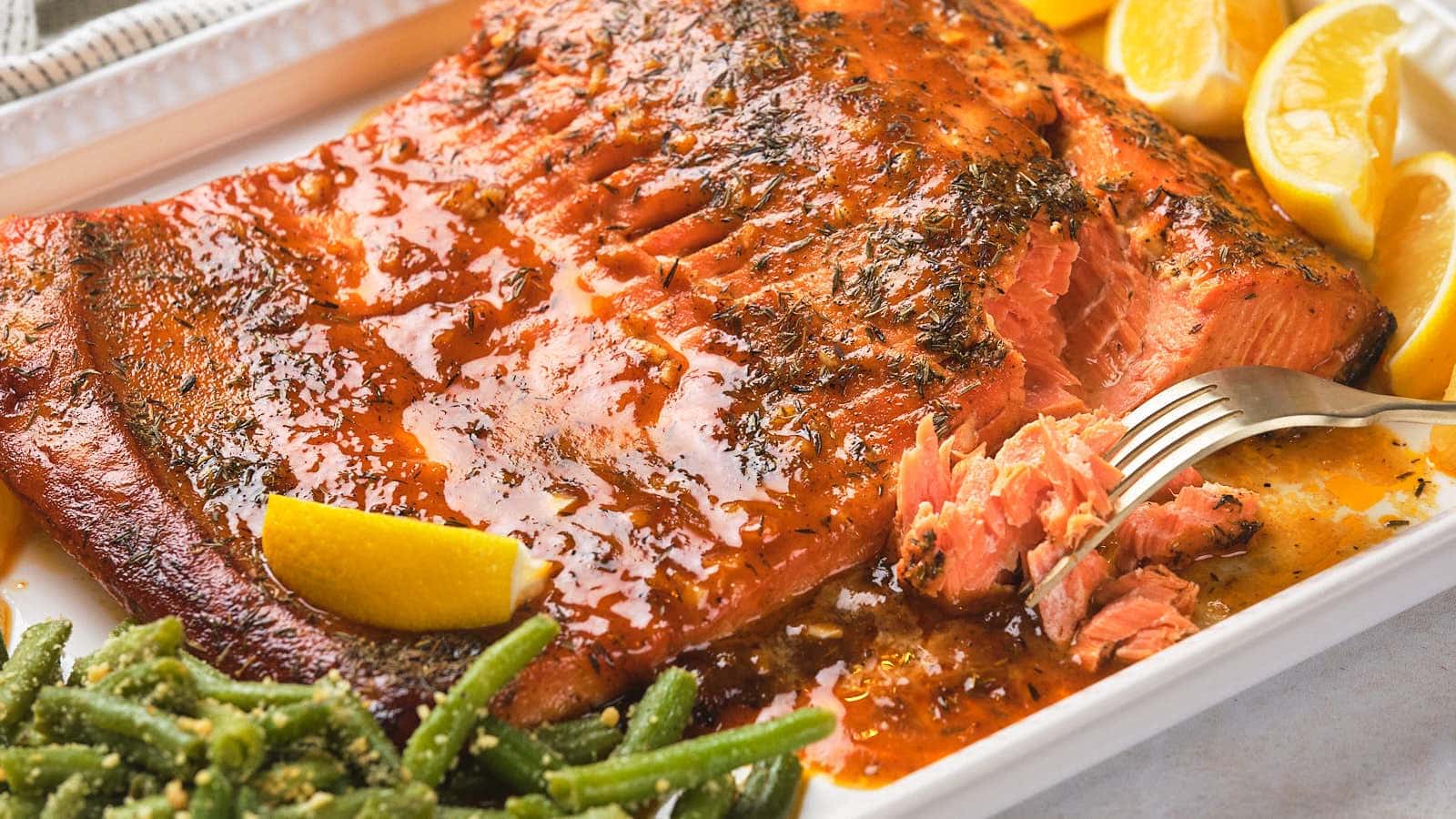 A baked salmon fillet glazed with sauce is served on a white platter with lemon wedges and green beans on the side. A fork holds a piece of the salmon.