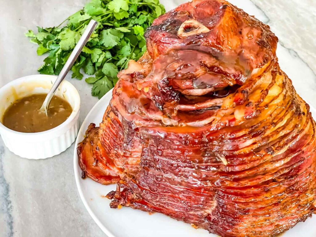 A spiral-sliced glazed ham sits on a white plate next to a bunch of cilantro and a small bowl of sauce with a spoon.