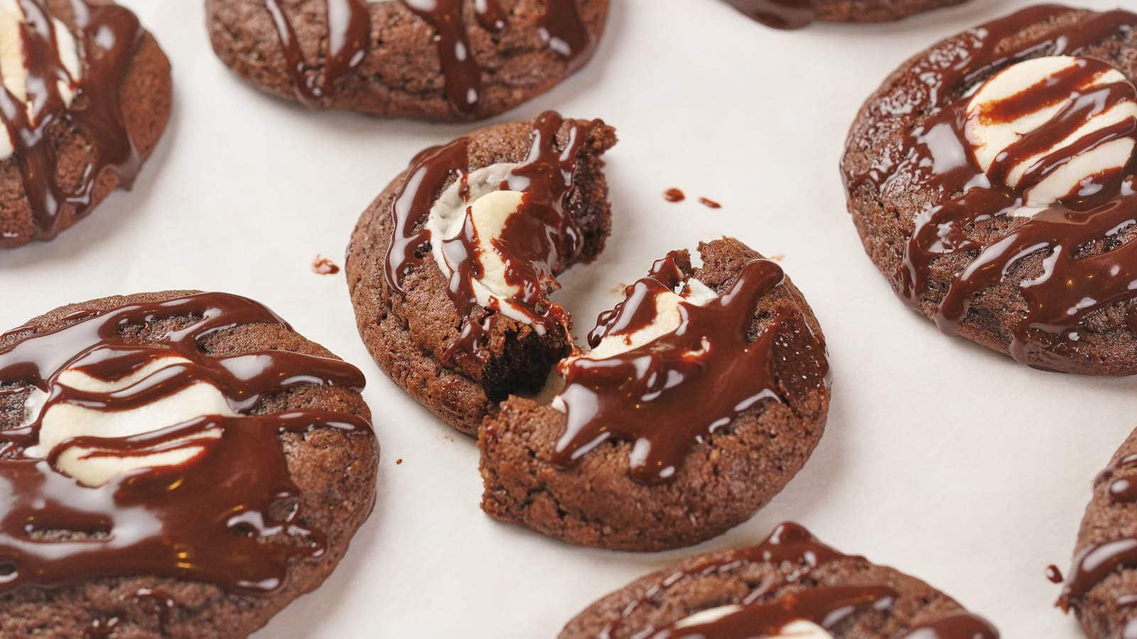 Chocolate cookies with a white swirl center, drizzled with chocolate, are arranged on a white surface; one cookie is broken in half.