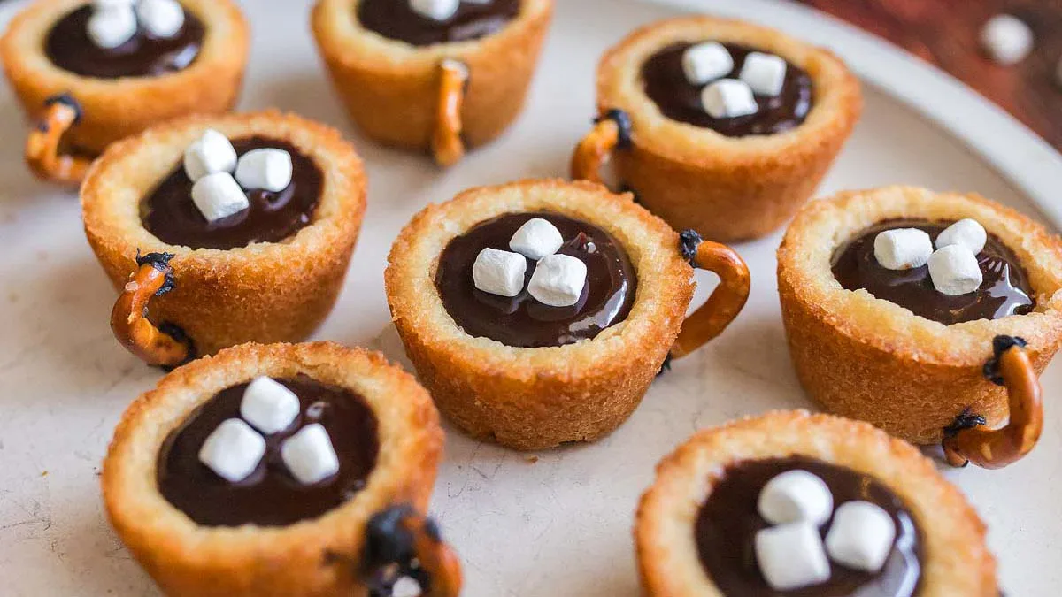 Mini cookie cups filled with chocolate and topped with mini marshmallows, designed to look like mugs, with pretzel piece handles, arranged on a white plate.