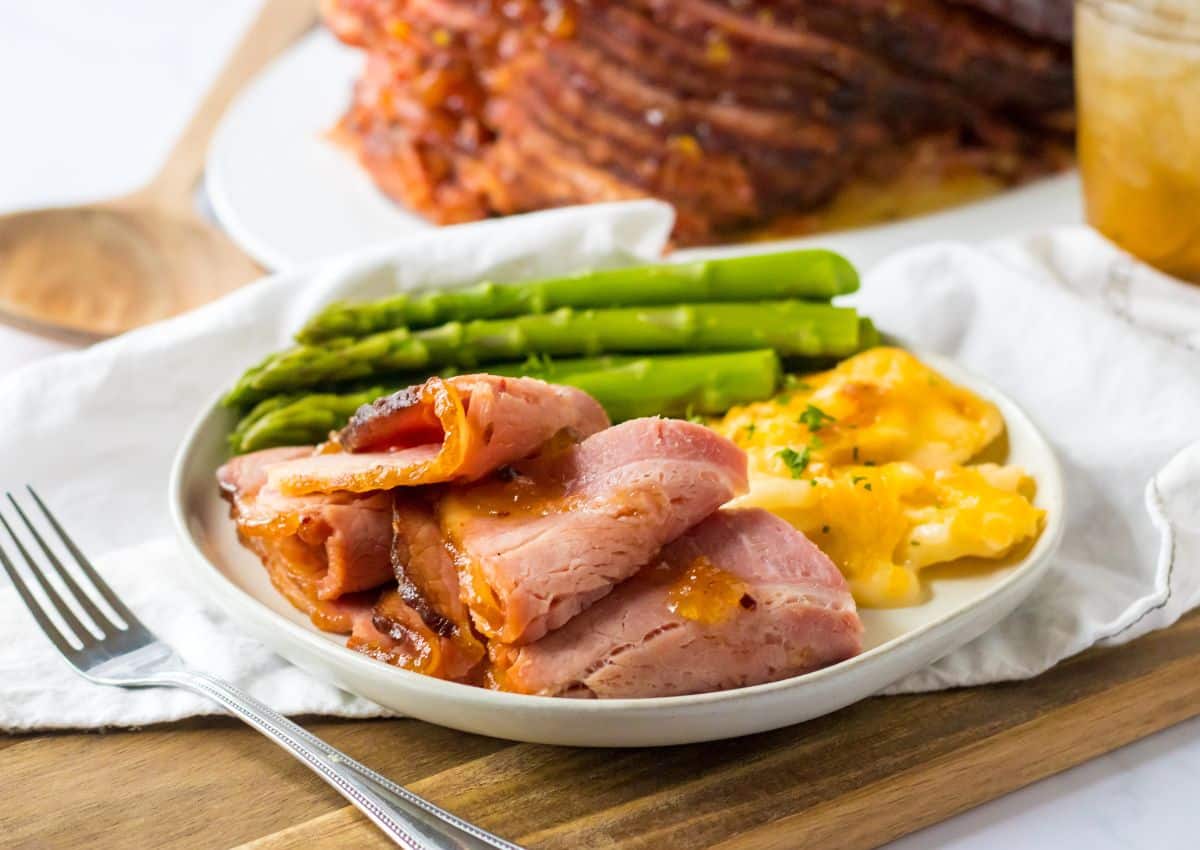 A plate with sliced ham, asparagus spears, and cheesy scalloped potatoes, set on a wooden board with a fork beside it.