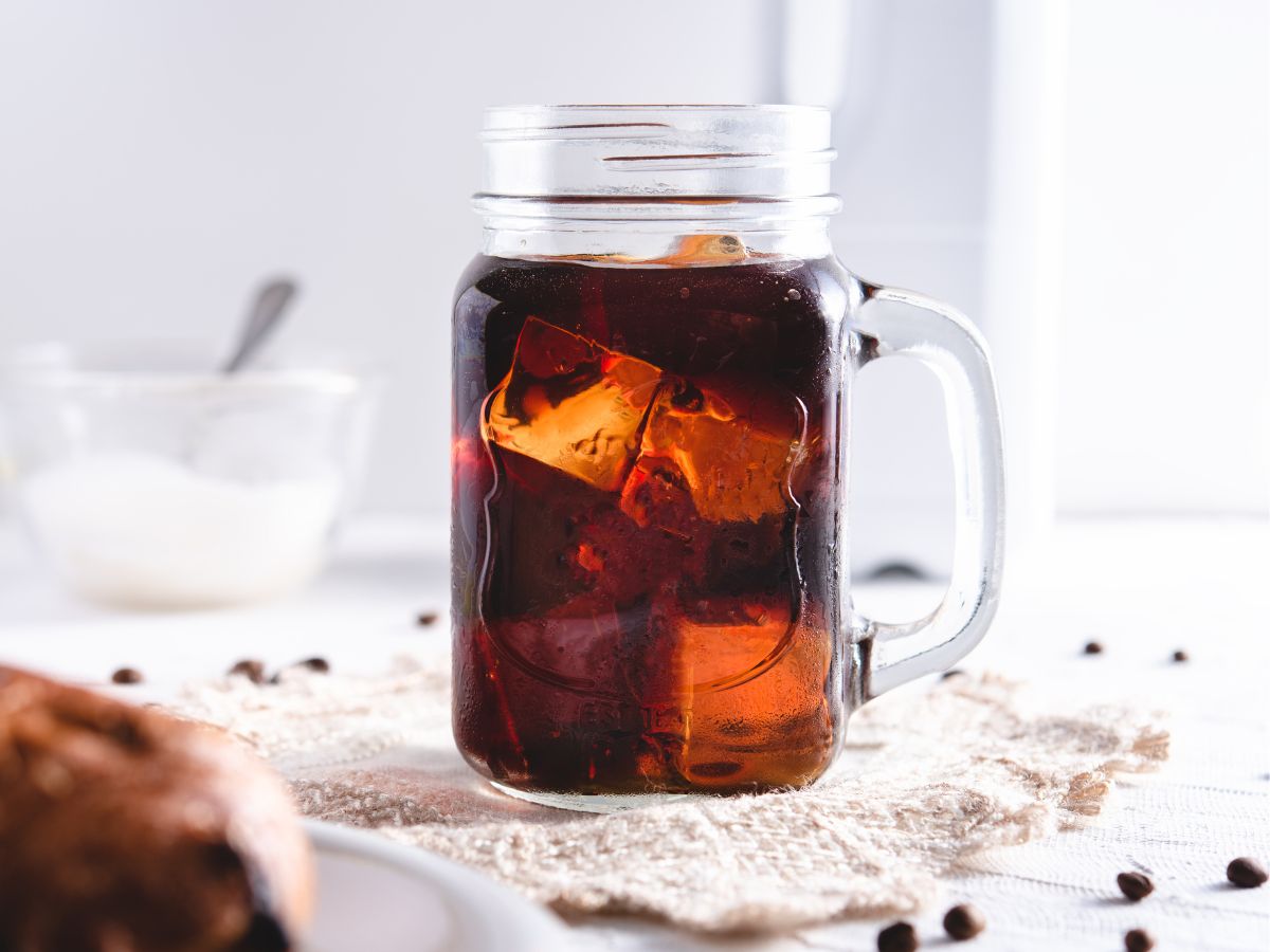 A glass mason jar filled with iced black coffee sits on a lace doily, with a blurred pastry and a bowl in the background.