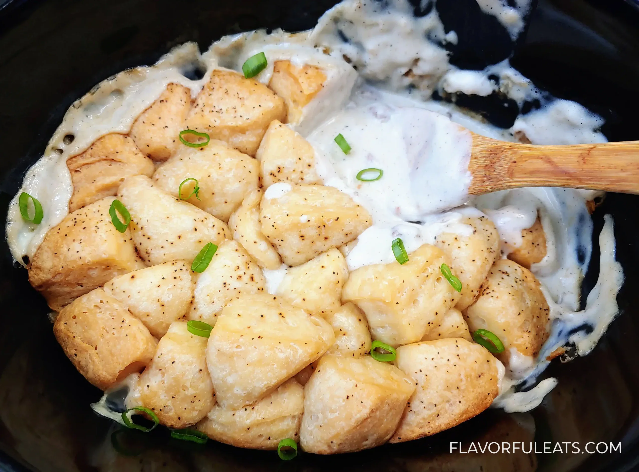 Baked biscuit pieces topped with sliced green onions in a creamy white sauce, being stirred with a wooden spoon in a black dish.