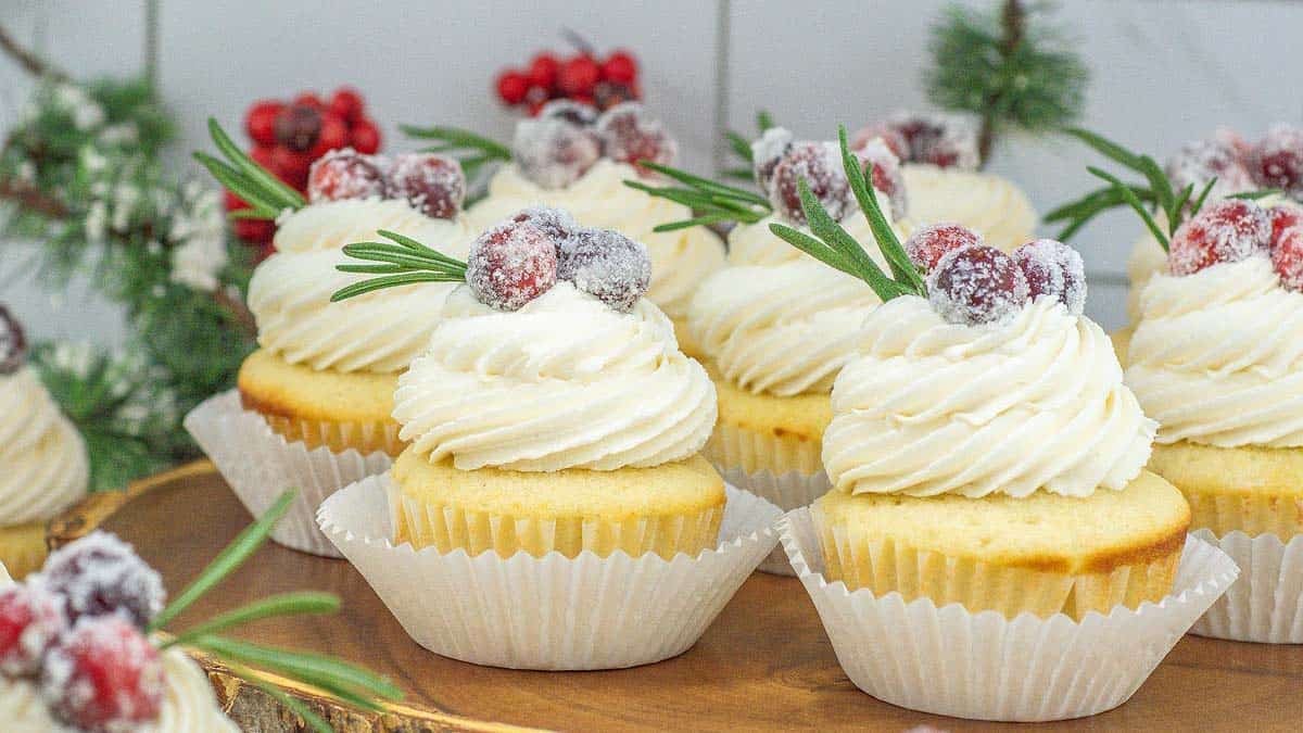 Vanilla cupcakes with swirled white frosting, topped with sugared cranberries and rosemary sprigs, arranged on a wooden platter with holiday greenery in the background.