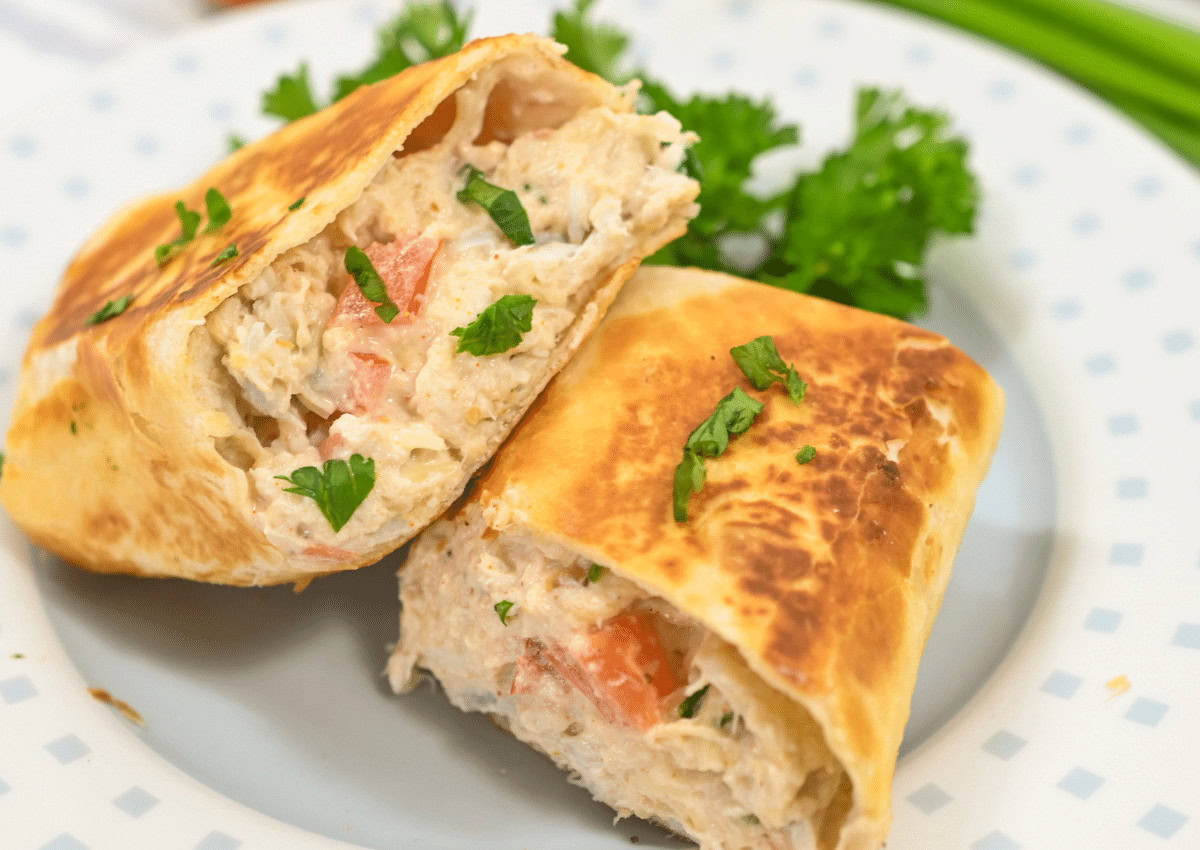 A toasted wrap cut in half, filled with a creamy chicken and vegetable mixture, garnished with chopped parsley, served on a white plate.