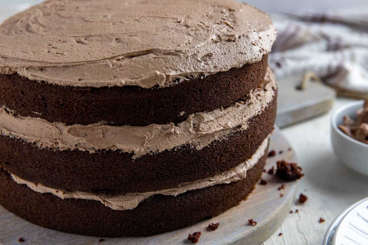 Three-layer chocolate cake with chocolate frosting between each layer and on top, displayed on a wooden board with a bowl of frosting in the background.