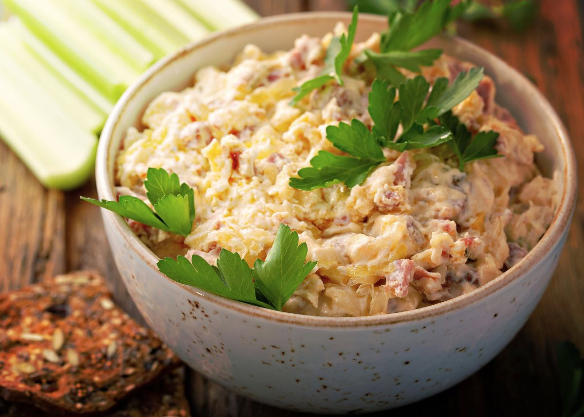 A bowl of creamy dip topped with fresh parsley, placed on a wooden surface with crackers and celery sticks beside it.