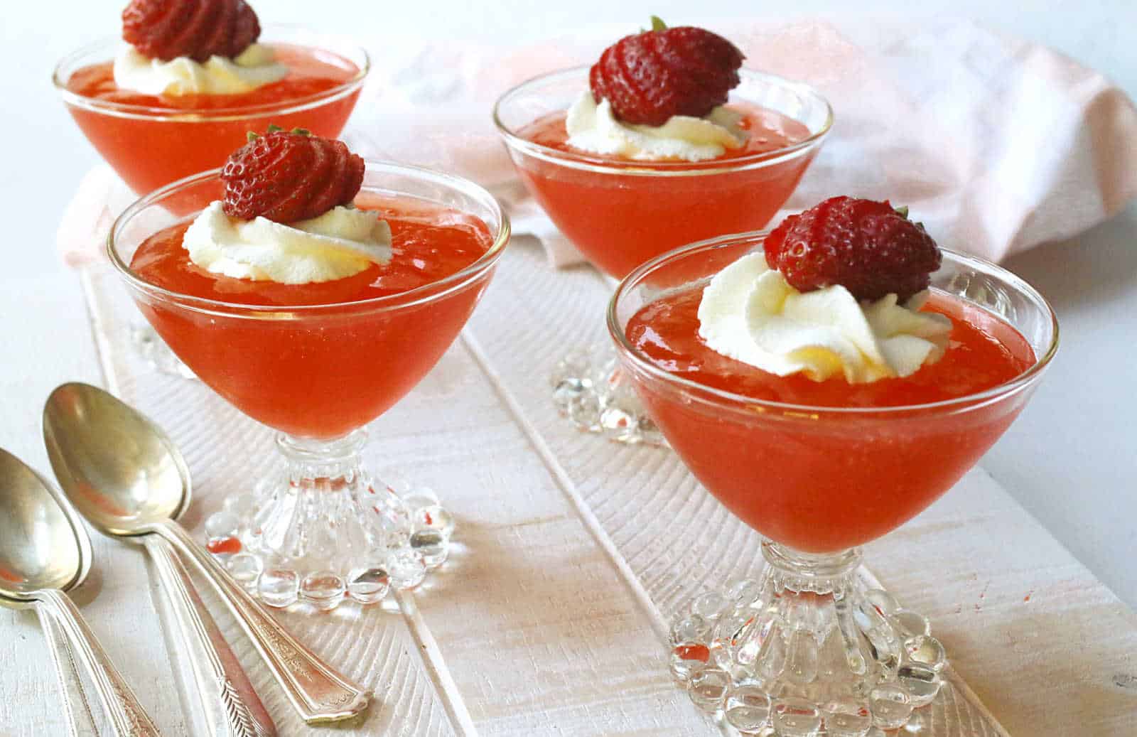 Four glass dessert cups filled with strawberry jelly, topped with whipped cream and a halved strawberry, placed on a white surface with three silver spoons beside them.