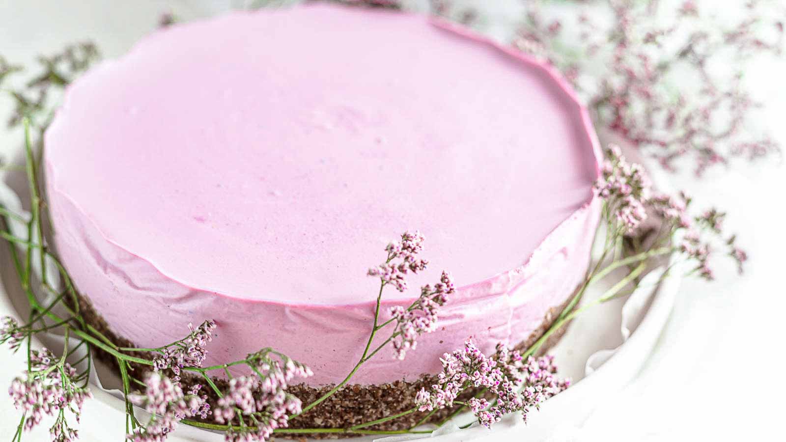 A round cake with a smooth pink frosting and a brown base, surrounded by small sprigs of pink flowers on a white plate.