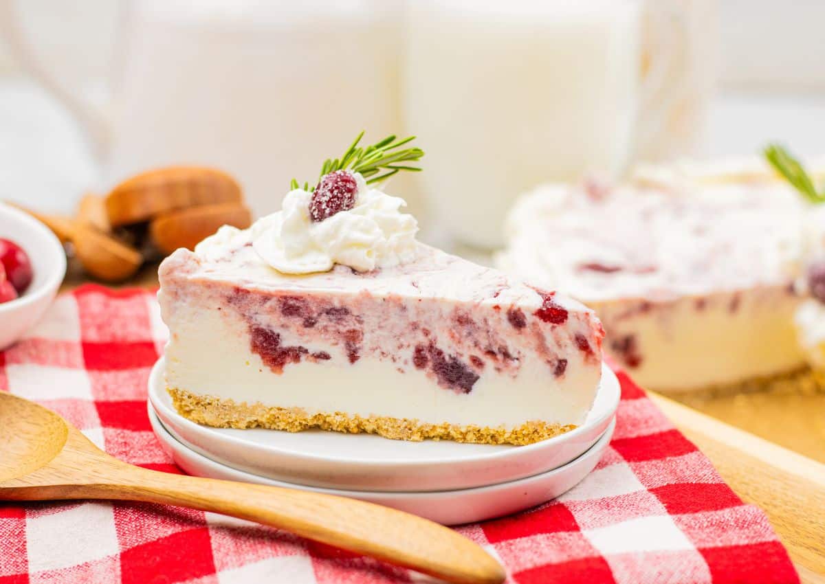 A slice of berry swirl cheesecake with a graham cracker crust, topped with whipped cream, a berry, and a sprig of rosemary, on a small plate beside a wooden spoon.