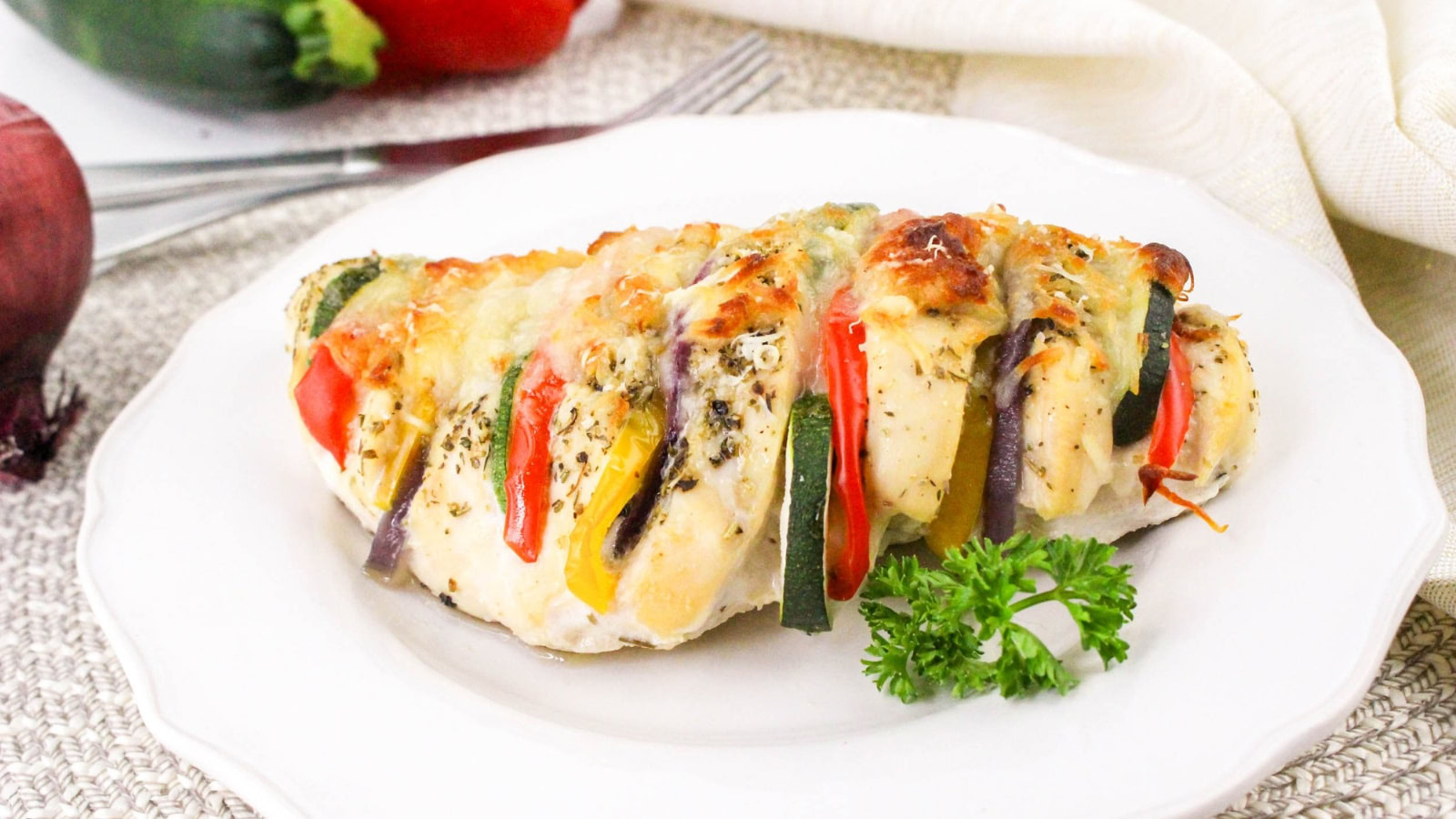 A stuffed chicken breast with sliced bell peppers, zucchini, and melted cheese, garnished with parsley, served on a white plate.