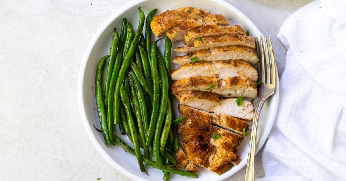 A white plate with sliced grilled chicken breast and cooked green beans, with a fork on the right side.