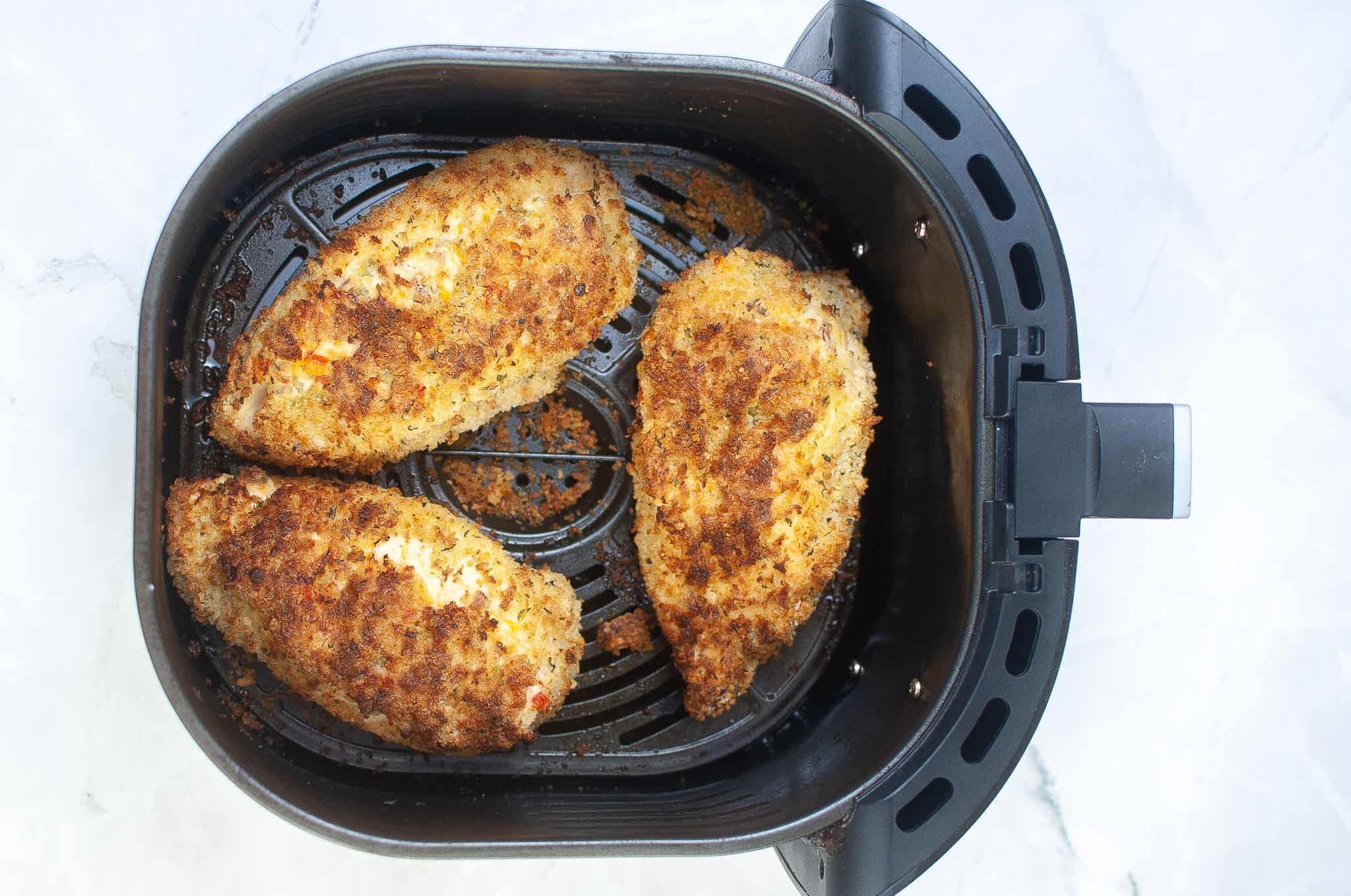 Three breaded chicken breasts are cooked and resting inside the basket of a black air fryer on a white surface.