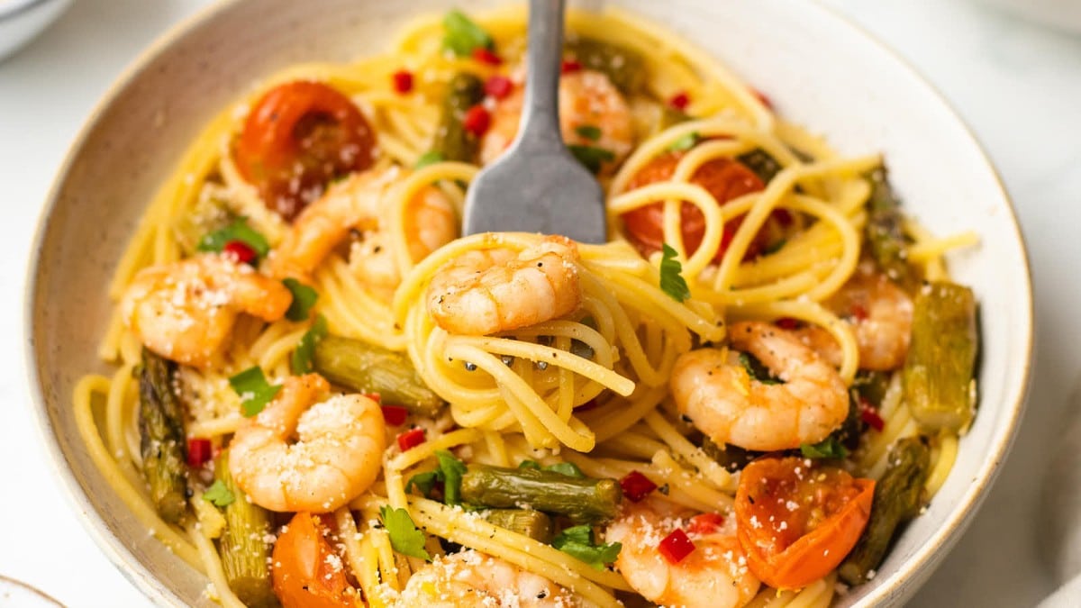 A bowl of spaghetti with shrimp, cherry tomatoes, asparagus, parsley, and grated cheese, with a fork twirling some pasta.