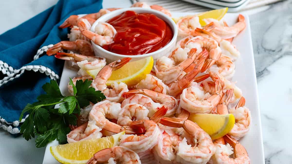 A platter of cooked shrimp is arranged around a small bowl of cocktail sauce, garnished with lemon wedges and parsley.