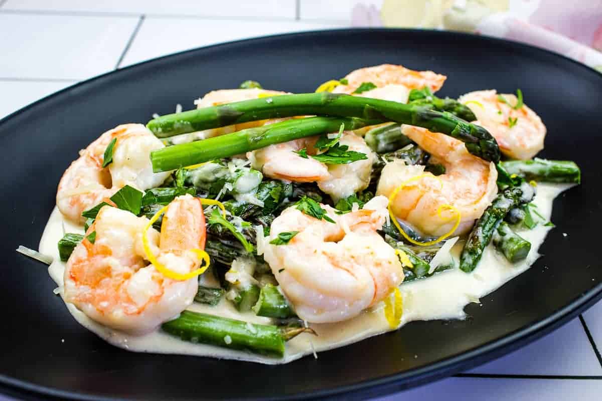 Shrimp and asparagus served on a black plate with creamy white sauce, garnished with lemon zest and parsley.