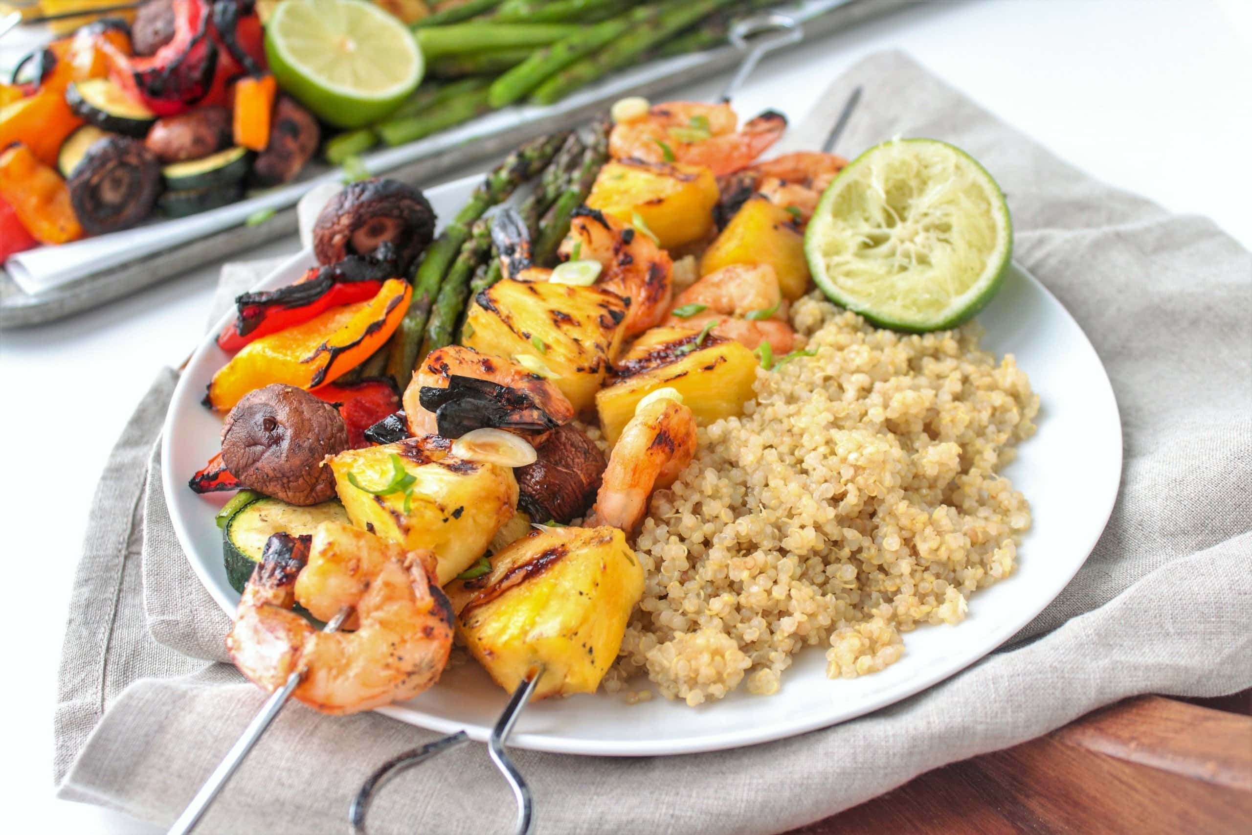 Grilled shrimp and vegetable skewers with pineapple served alongside cooked quinoa, grilled asparagus, and a halved lime on a white plate.