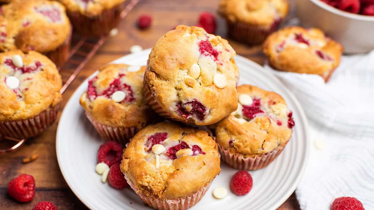 A plate of raspberry and white chocolate chip muffins is surrounded by fresh raspberries, with more muffins and a bowl of raspberries in the background.