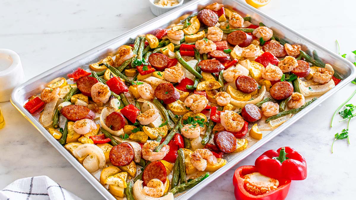 A sheet pan filled with cooked shrimp, sliced sausage, bell peppers, onions, and squash, all seasoned and roasted, rests on a white surface with fresh vegetables nearby.
