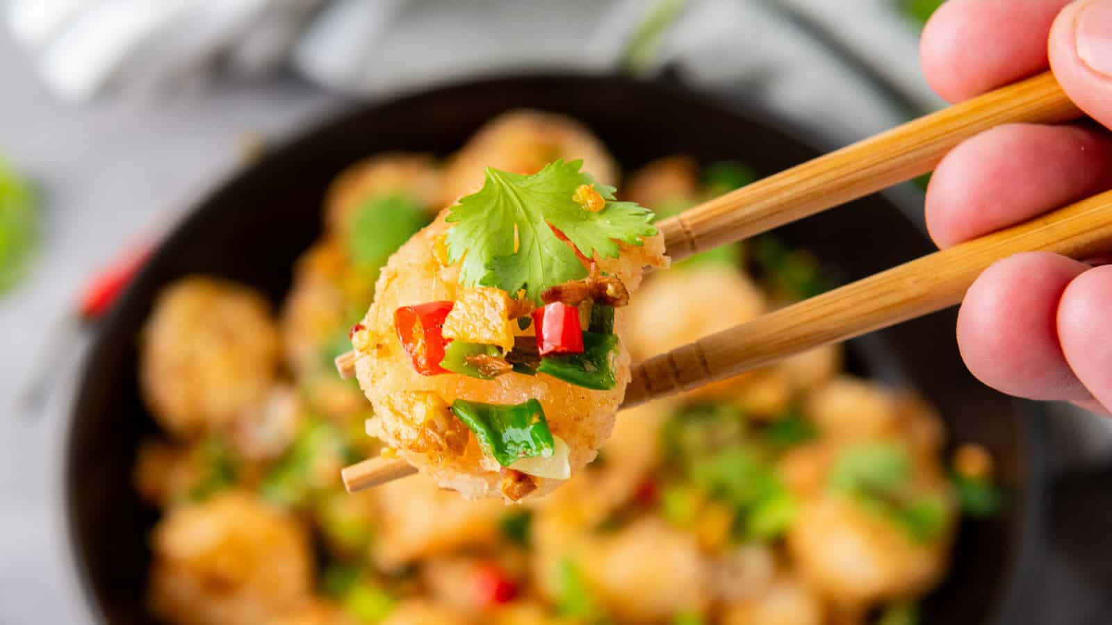 A hand holds chopsticks with a shrimp topped with cilantro, chopped red and green peppers, and seasoning, with more shrimp on a plate in the background.